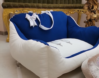 May include: A white and blue pet bed with a white strap and a pink bow. The bed has a white, padded base and a blue fabric top with a white strap. The bed is sitting on a wooden floor.