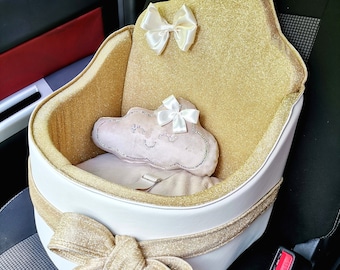 May include: A white car seat with a gold glitter fabric and a gold ribbon bow. The seat has a white cloud-shaped pillow with pink glitter and a white bow. The seat is attached to the car seat with a white strap.