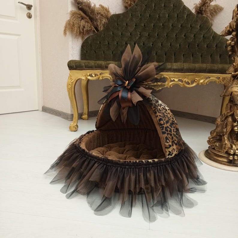 May include: A brown and black leopard print pet bed with a black tulle skirt and a brown bow on top. The bed is in a room with a gold and green chair.