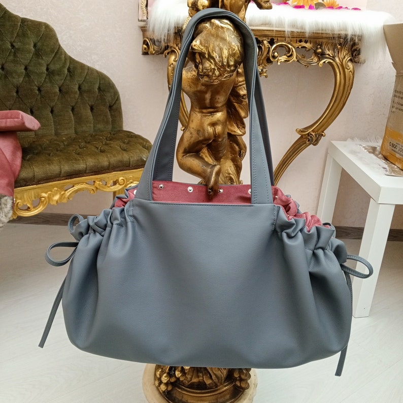May include: A grey, slouchy tote bag with a pink lining and a drawstring closure. The bag has a relaxed, casual look and is made of a soft, supple material.