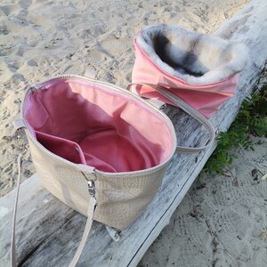 May include: A beige faux leather tote bag with a pink lining and a top zipper closure. The bag has a long strap and is shown open with a pink faux fur lining inside.