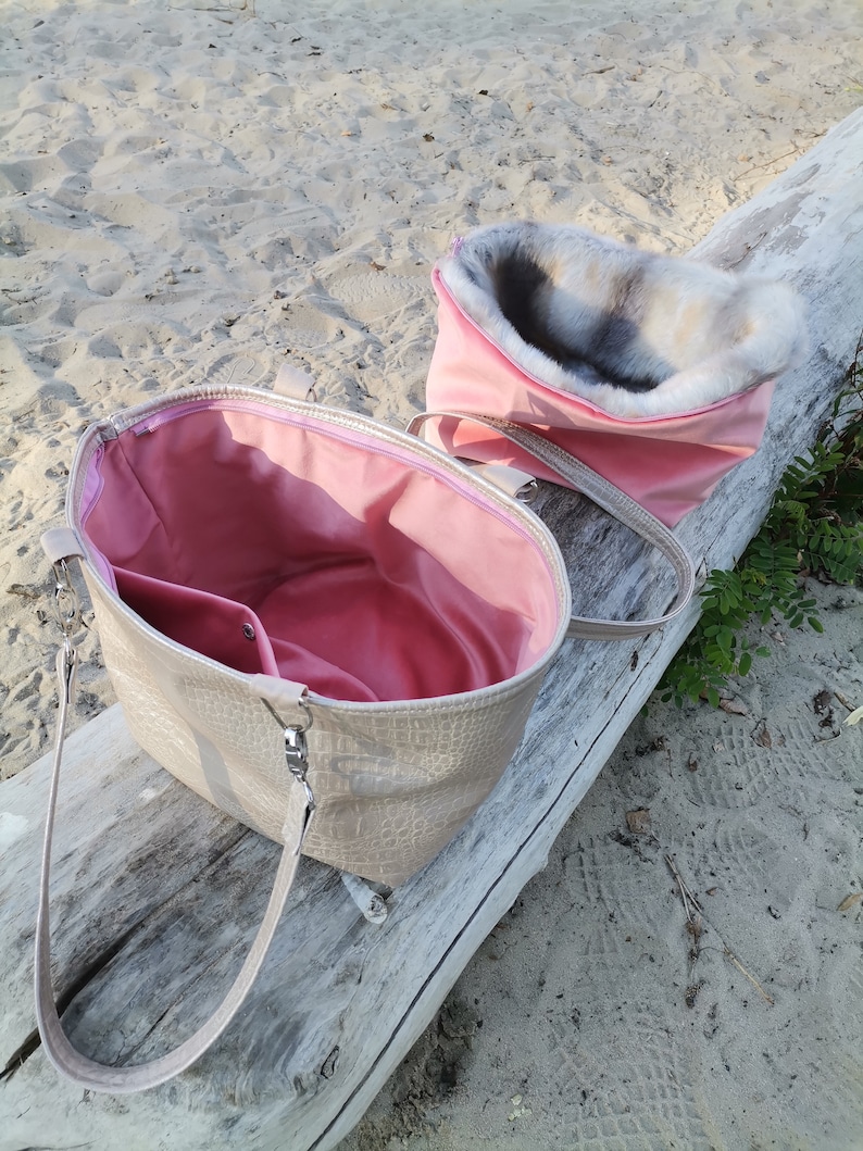 May include: A beige faux leather tote bag with a pink lining and a top zipper closure. The bag has a long strap and is shown open with a pink faux fur lining inside.