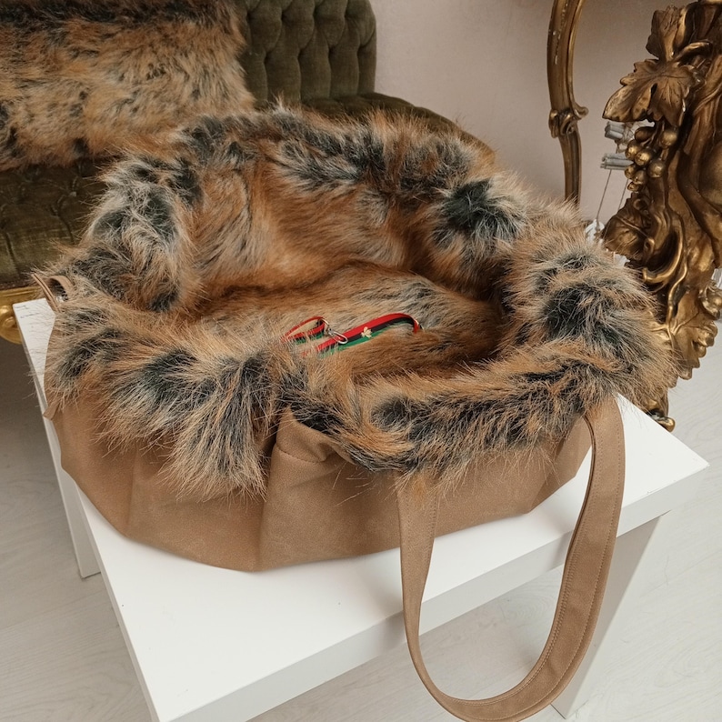 Pu&ograve; includere: Una borsa tote in pelliccia sintetica marrone con una grande fodera in pelliccia sintetica marrone e nera soffice. La borsa ha due manici marroni ed &egrave; appoggiata su una superficie bianca.