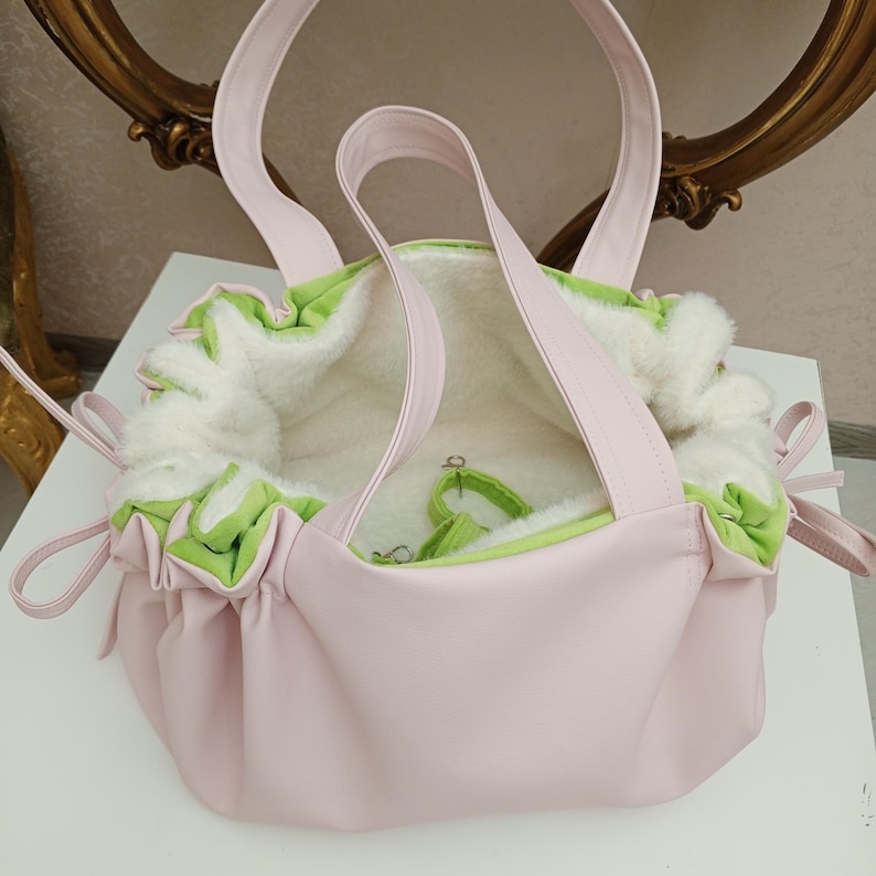 May include: A light pink tote bag with a soft, white interior and green trim. The bag has two handles and a drawstring closure. The exterior is a smooth, solid pink, and the interior is lined with a fluffy, white material.