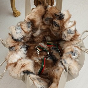 K&ouml;nnte beinhalten: Eine beige Tasche mit Kunstfellfutter und einem langen Trageriemen. Die Tasche ist offen und das Futter ist sichtbar. Das Futter ist aus braunem und wei&szlig;em Kunstfell.