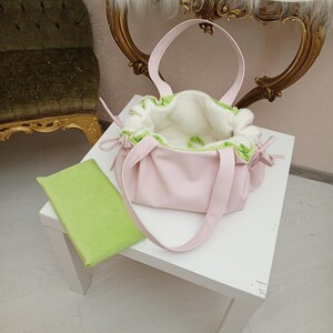 May include: A pink handbag with a white interior and green trim sits on a white table. The bag has two handles and is next to a green rectangular item. The background includes a gold-framed mirror and a green chair.