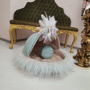 May include: A light blue and beige pet bed with a ruffled trim and a decorative bow. The bed has a round cushion with the name "Anna" embroidered on it.