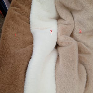 May include: Close-up of three faux fur fabrics in shades of brown, white, and tan. The textures are soft and plush, ideal for crafting or fashion projects. The image highlights the color variations and the tactile quality of the materials.