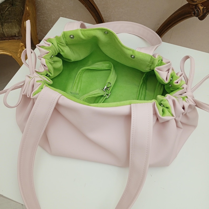 May include: A light pink tote bag with a bright green interior. The bag has two handles and a drawstring closure. The interior includes a small, detachable green strap with a silver clasp. The bag's exterior is a smooth, solid color.