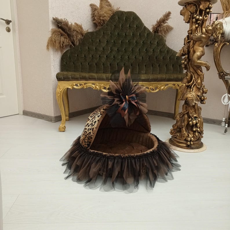 May include: A brown and black leopard print pet bed with a black tulle skirt and a brown satin bow. The bed is sitting on a white floor in front of a gold and brown ornate stand.