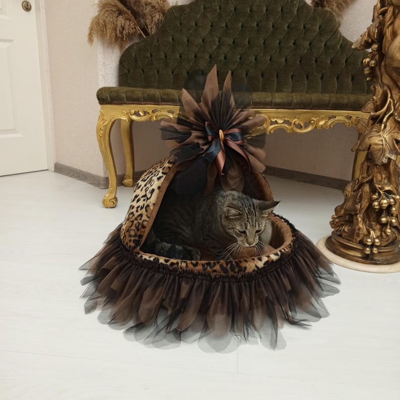May include: A brown and black leopard print cat bed with a brown tulle skirt and a brown bow on top. A gray tabby cat is inside the bed.