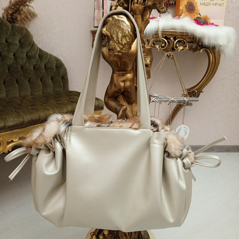 K&ouml;nnte beinhalten: Eine hellbeige Ledertasche mit Kunstfellbesatz. Die Tasche hat zwei obere Henkel und einen Kordelverschluss.