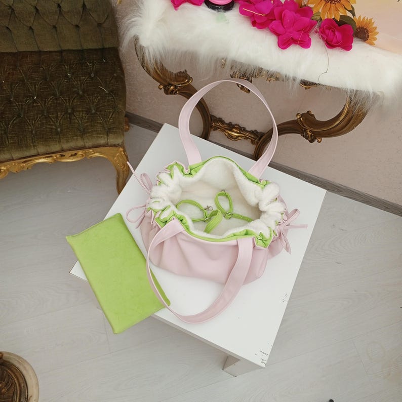 May include: A light pink handbag with matching handles and a drawstring closure. The interior is white with green trim. A lime green rectangular item rests beside the bag. The bag is on a white surface.