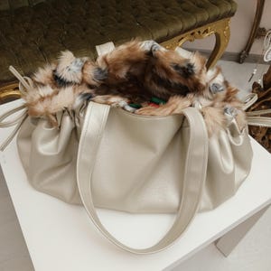 K&ouml;nnte beinhalten: Eine goldfarbene Kunstleder-Tragetasche mit einem flauschigen braunen und wei&szlig;en Kunstfellfutter. Die Tasche hat zwei Henkel und ein gerafftes Design.