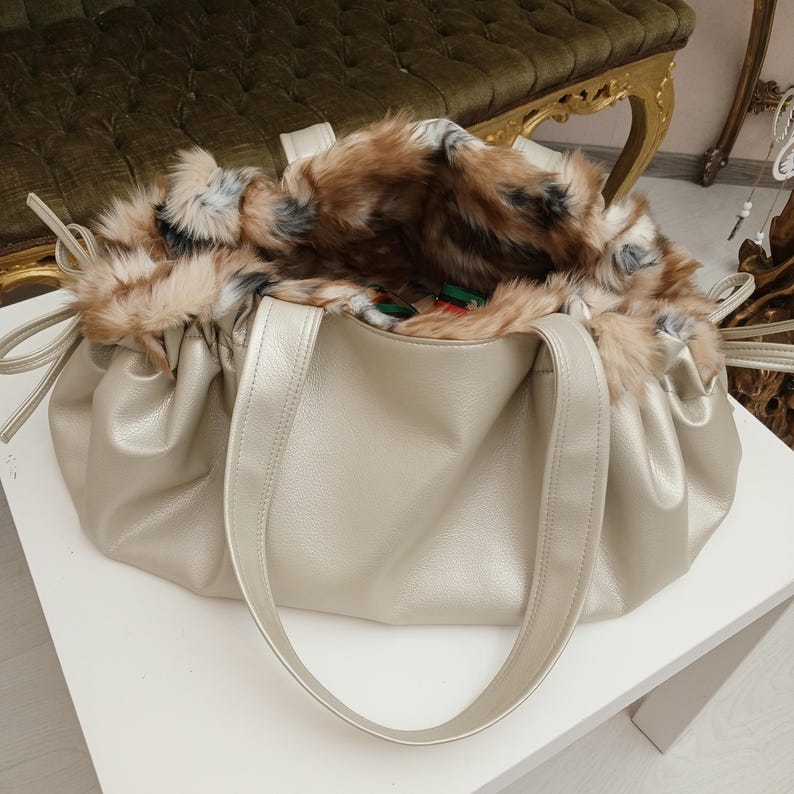 K&ouml;nnte beinhalten: Eine goldfarbene Kunstleder-Tragetasche mit einem flauschigen braunen und wei&szlig;en Kunstfellfutter. Die Tasche hat zwei Henkel und ein gerafftes Design.