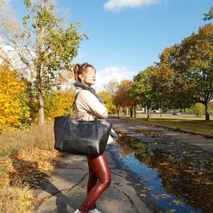 May include: A woman wearing a beige jacket, a black scarf, and red leather pants is carrying a large black tote bag with a crocodile-embossed design. She is standing on a paved path in a park with autumn leaves on the ground.