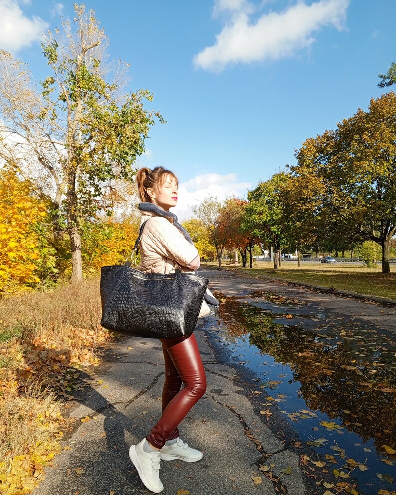 May include: A woman wearing a beige jacket, a black scarf, and red leather pants is carrying a large black tote bag with a crocodile-embossed design. She is standing on a paved path in a park with autumn leaves on the ground.