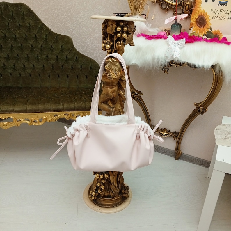 May include: A pale pink faux leather handbag with a white faux fur lining and a drawstring closure. The bag is sitting on a gold stand.
