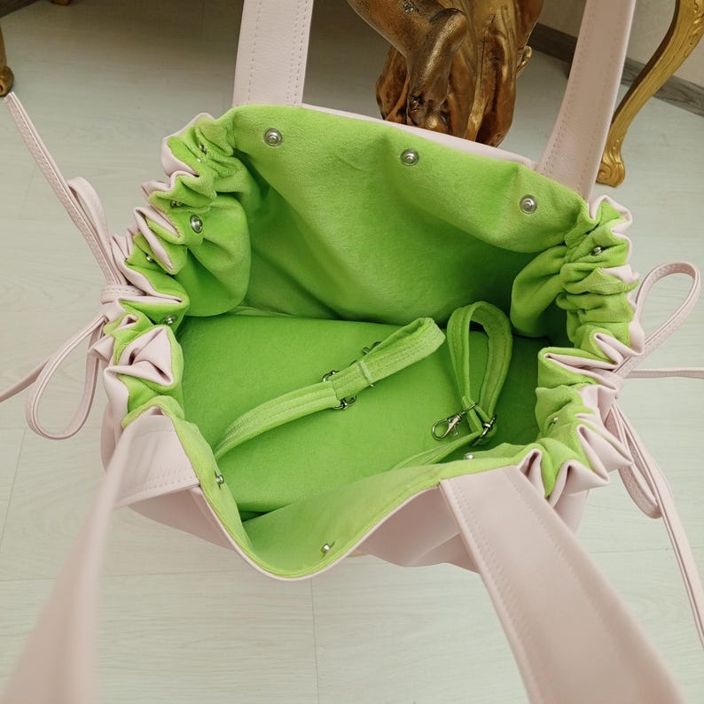 May include: A light pink handbag with a drawstring closure and a bright green interior. The bag has a soft, velvety texture and a shoulder strap. The interior features a small, adjustable strap with a silver clasp. The bag's design is simple and elegant.