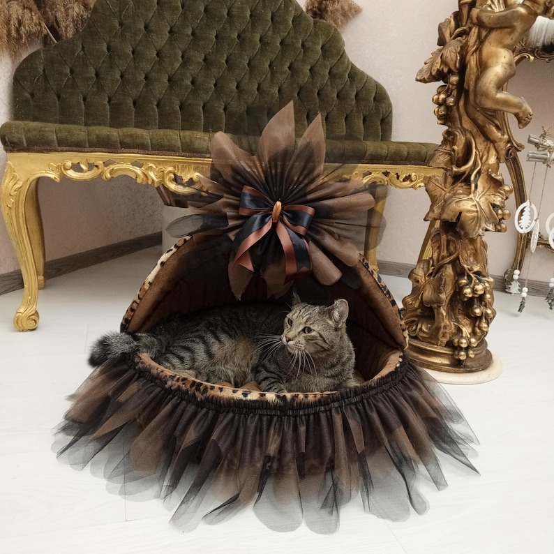 May include: A brown and black cat bed with a tulle skirt and a brown bow. The bed is made of a soft, plush material and has a comfortable, cushioned interior. The cat is lying in the bed, looking at the camera.