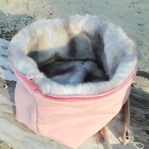 May include: A pink fabric pet bed with a faux fur lining. The bed has a zipper closure and a brown strap.