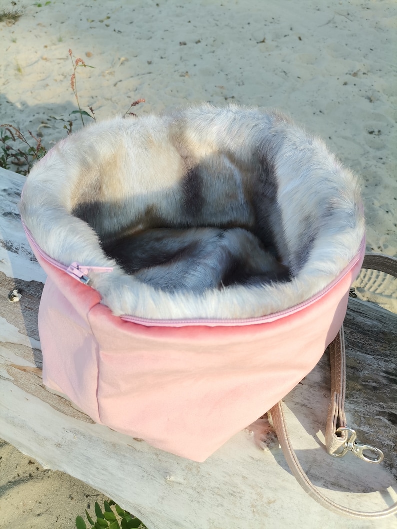 May include: A pink fabric pet bed with a faux fur lining. The bed has a zipper closure and a brown strap.