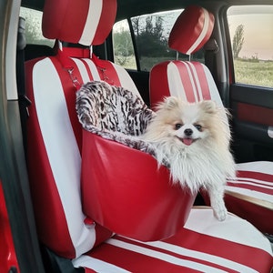 Puede incluir: Fundas de asiento de coche rojas y blancas con un asiento de coche para perros a juego. El asiento de coche para perros tiene un interior con estampado de leopardo y está sujeto al reposacabezas. Un perro pequeño, esponjoso y de color claro está sentado en el asiento del coche.