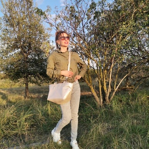 May include: A woman wearing a green shirt, gray jeans, and white sneakers is standing in a field. She is carrying a light-colored handbag with a strap over her shoulder. She is looking up and smiling.