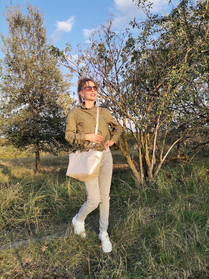 May include: A woman wearing a green shirt, gray jeans, and white sneakers is standing in a field. She is carrying a light-colored handbag with a strap over her shoulder. She is looking up and smiling.