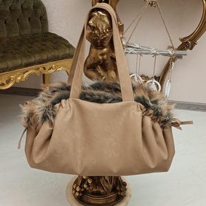 Pu&ograve; includere: Una borsa tote in similpelle color beige con un bordo in pelliccia. La borsa &egrave; appoggiata su un piedistallo dorato.