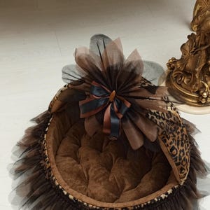 May include: A brown and black leopard print pet bed with a black tulle skirt and a brown and black bow. The bed has a brown velvet lining.