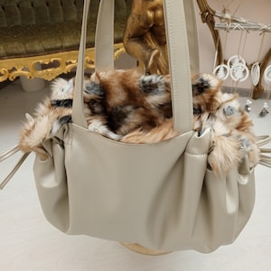 K&ouml;nnte beinhalten: Eine beige Ledertasche mit einem Futter aus Kunstfell. Die Tasche hat zwei obere Henkel und einen Kordelzugverschluss.