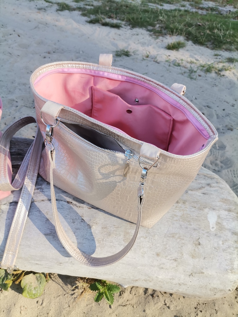 May include: A gold-colored tote bag with a pink lining and a zipper closure. The bag has a long strap and a shorter handle.