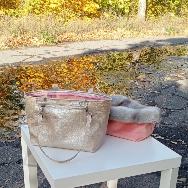 May include: A gold-colored tote bag with a crocodile-embossed design and a pink lining. The bag is sitting on a white table next to a pink faux fur bag.