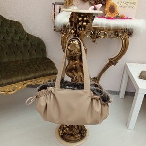 May include: A beige faux leather pet carrier with a brown faux fur trim. The carrier has a top handle and a drawstring closure. The carrier is sitting on a gold decorative stand.