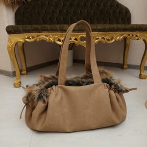 Pu&ograve; includere: Una borsa tote in pelle marrone con un bordo in pelliccia sintetica intorno alla parte superiore. La borsa ha due manici e una chiusura con coulisse.