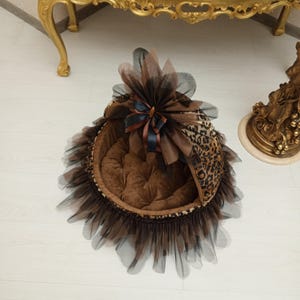 May include: A brown and black leopard print pet bed with a brown velvet cushion. The bed has a black tulle ruffle around the bottom and a black tulle canopy with a brown satin bow on top.