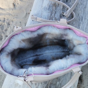 May include: A light beige tote bag with a pink zipper and a fluffy gray faux fur lining. The bag has a long strap and two shorter handles.