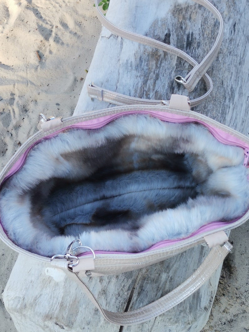May include: A light beige tote bag with a pink zipper and a fluffy gray faux fur lining. The bag has a long strap and two shorter handles.