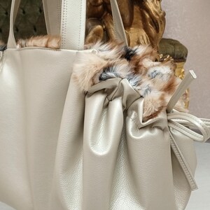 K&ouml;nnte beinhalten: Eine hellbeige Lederhandtasche mit Kordelzugverschluss und Pelzbesatz. Die Tasche hat zwei obere Henkel und einen langen Riemen.