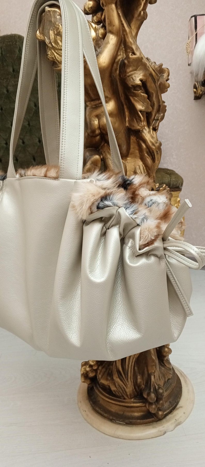 K&ouml;nnte beinhalten: Eine hellbeige Lederhandtasche mit Kordelzugverschluss und Pelzbesatz. Die Tasche hat zwei obere Henkel und einen langen Riemen.