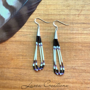 May include: A pair of dangle earrings with black, white, and purple beads, and white feathers. The earrings are silver and have a hook closure.