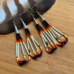 May include: A pair of long, beaded earrings with black, red, yellow, and white beads. The earrings are made with bone and feathers and have silver hooks.