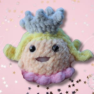 May include: A handmade, plush doll in the shape of a princess. The doll features a light peach face, yellow hair, and a gray crown. The doll has black eyes and a pink and peach body. The background is pink with star-shaped confetti.