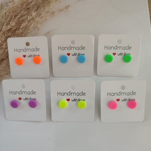 Op de afbeelding: Zes paar handgemaakte oorbellen gepresenteerd op witte kaartjes. De oorbellen zijn klein en rond, in verschillende felle kleuren, waaronder oranje, blauw, groen, geel, paars en roze. Elk kaartje heeft de tekst "Handmade with love" erop gedrukt.