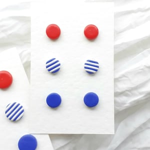 May include: Six pairs of small, round earrings displayed on a white card. The earrings are red, blue, and blue with white stripes.