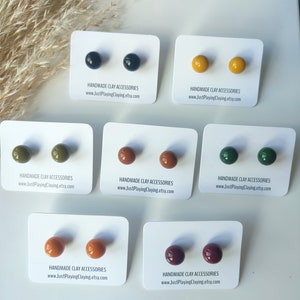 Peut inclure: Six paires de boucles d'oreilles en argile faites à la main dans différentes couleurs, notamment noir, jaune, vert, marron et rouge. Chaque paire est présentée sur une carte blanche avec le texte "HANDMADE CLAY ACCESSORIES" et "www.JustPlayingClaying.etsy.com".