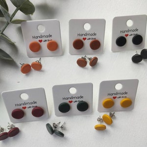 Può includere: Sei paia di orecchini a bottone fatti a mano su carte bianche. Gli orecchini sono di vari colori, tra cui rosso, arancione, marrone, nero, verde e giallo. Ogni carta ha il testo "Handmade with love" stampato su di essa.