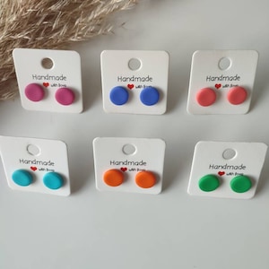 May include: Six pairs of handmade polymer clay stud earrings displayed on white cards. The earrings are in various colours, including pink, blue, orange, and green. Each card has the text "Handmade with love" printed on it.