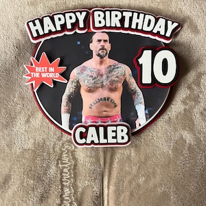 May include: A personalized birthday cake topper featuring a photo of a man with tattoos, the text "HAPPY BIRTHDAY 10 CALEB", and a "BEST IN THE WORLD" starburst. The topper is round with a black background and red and white accents.